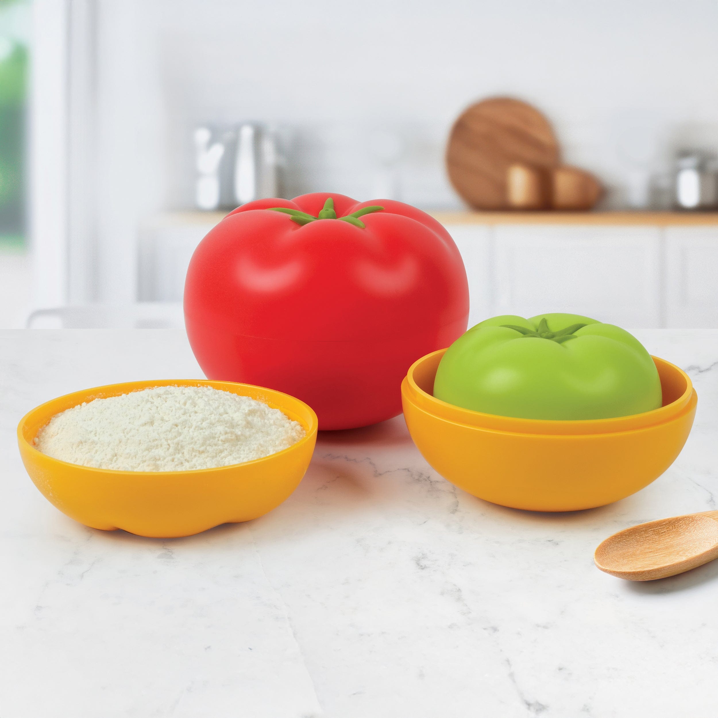 Red, yellow, and green tomato-shaped measuring cups, measuring flour on a kitchen counter.