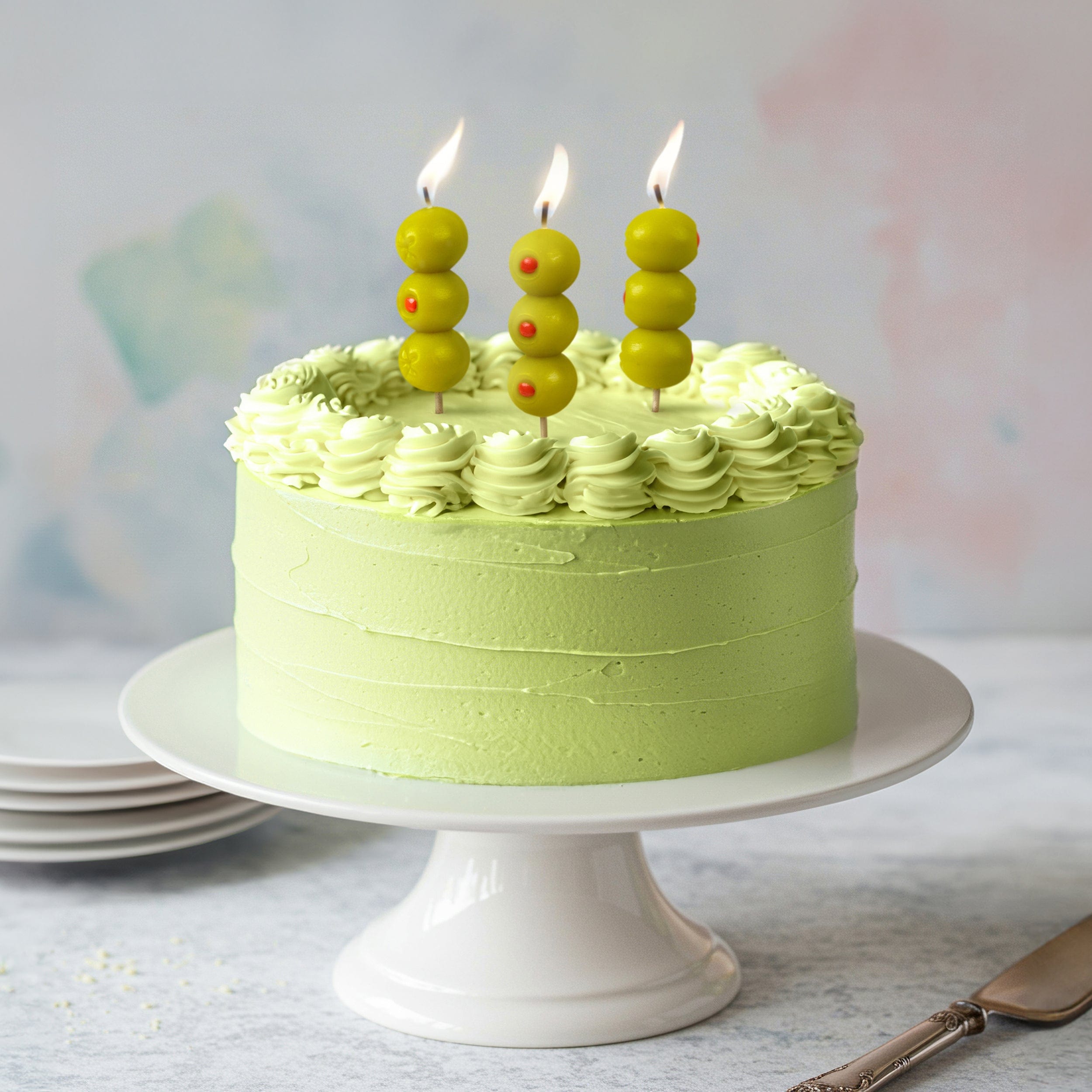 Green cake with birthday candles that look like cocktail olives on on a white cake stand against a light background