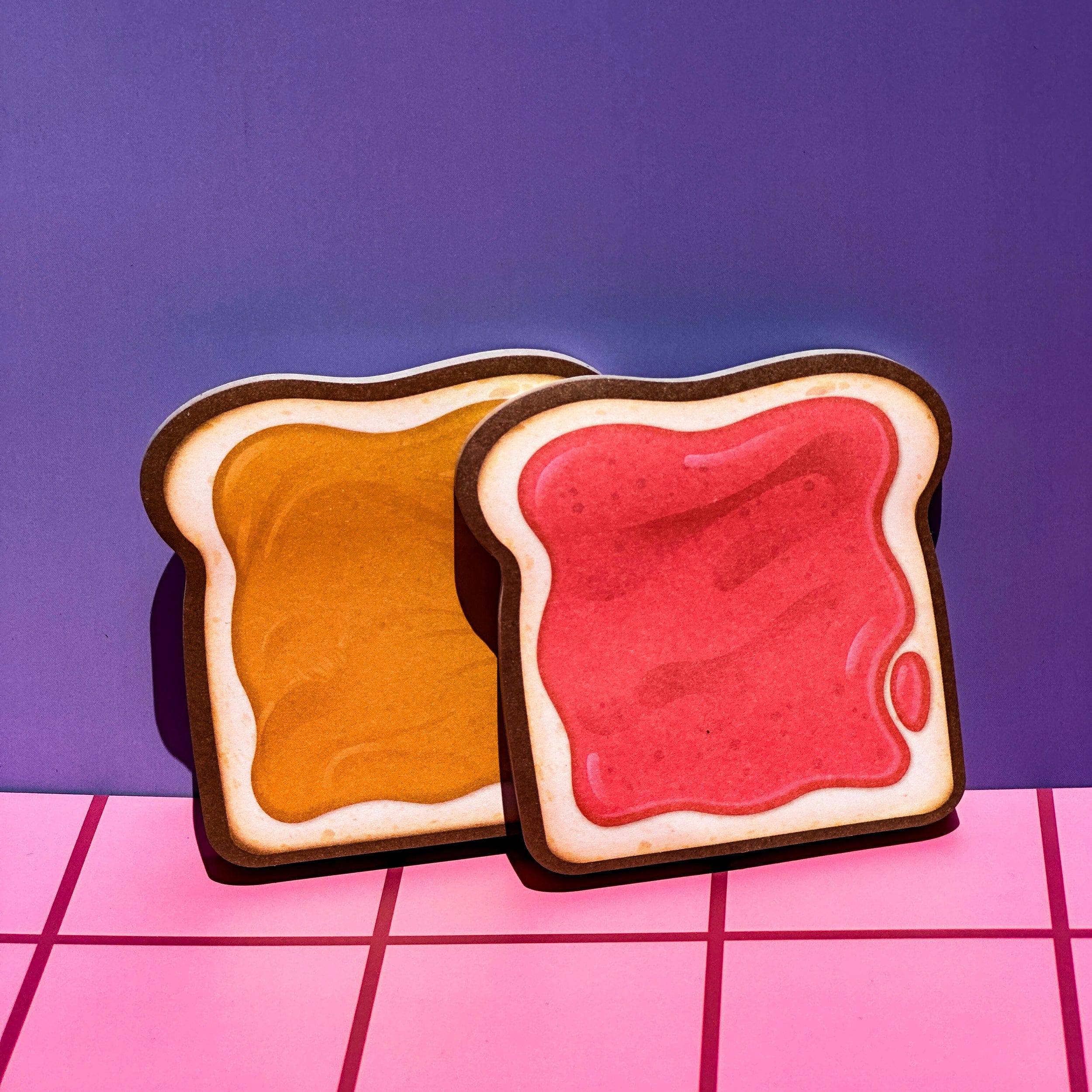 Two sticky note pads with with peanut butter and jelly on a pink and purple grid background