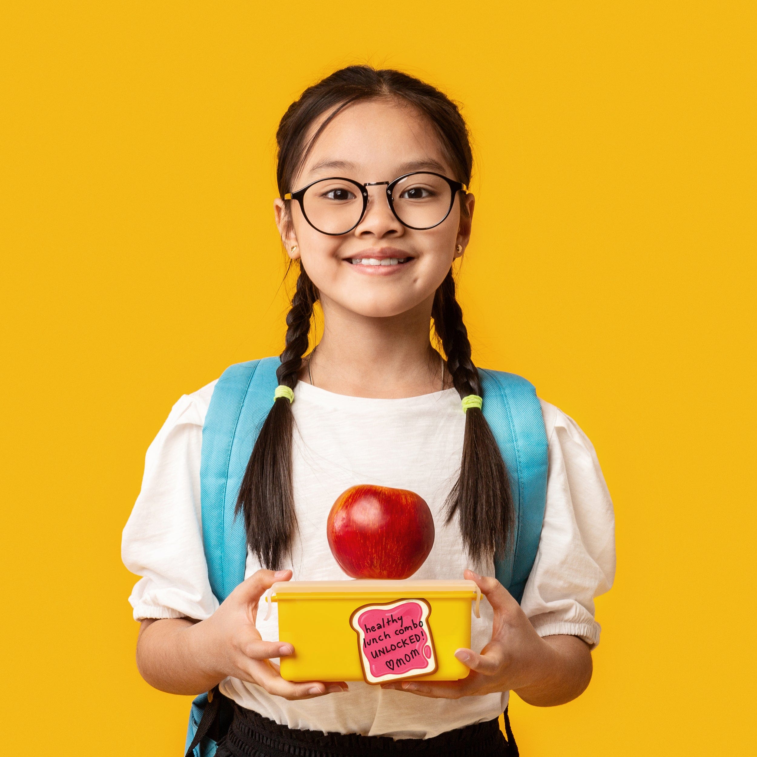 Young girl with glasses holding a lunchbox with an apple and a jelly sticky note on a yellow background