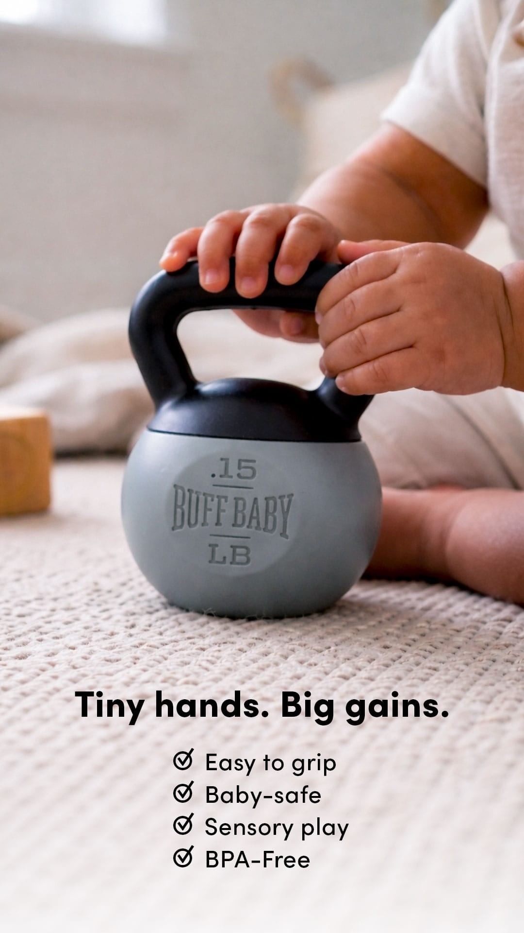 Child's hand holding a small gray kettlebell rattle with 'Buff Baby' branding on a light surface.