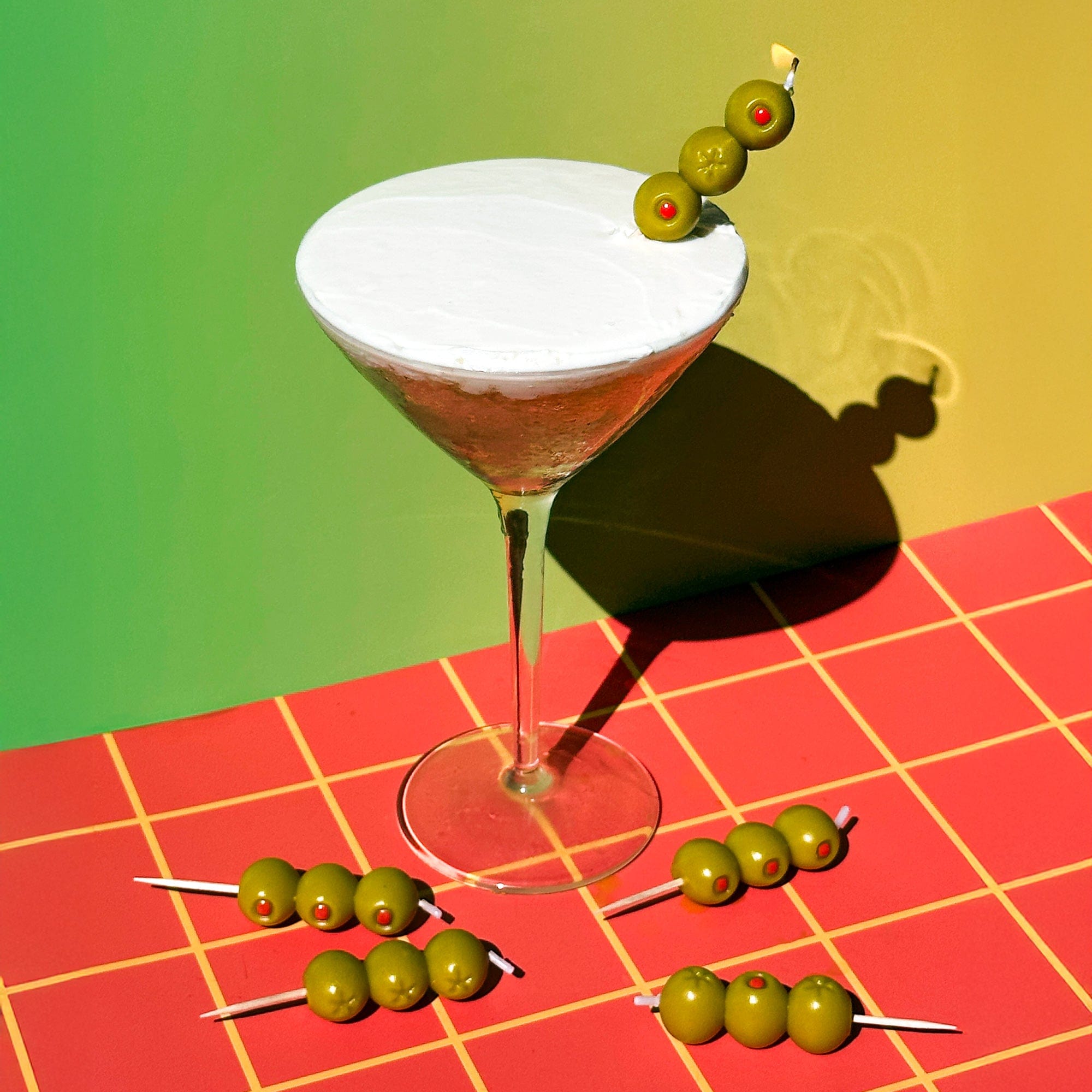 Cocktail cake with a birthday candle that looks like cocktail olives on a red and yellow checkered tablecloth and green wall.