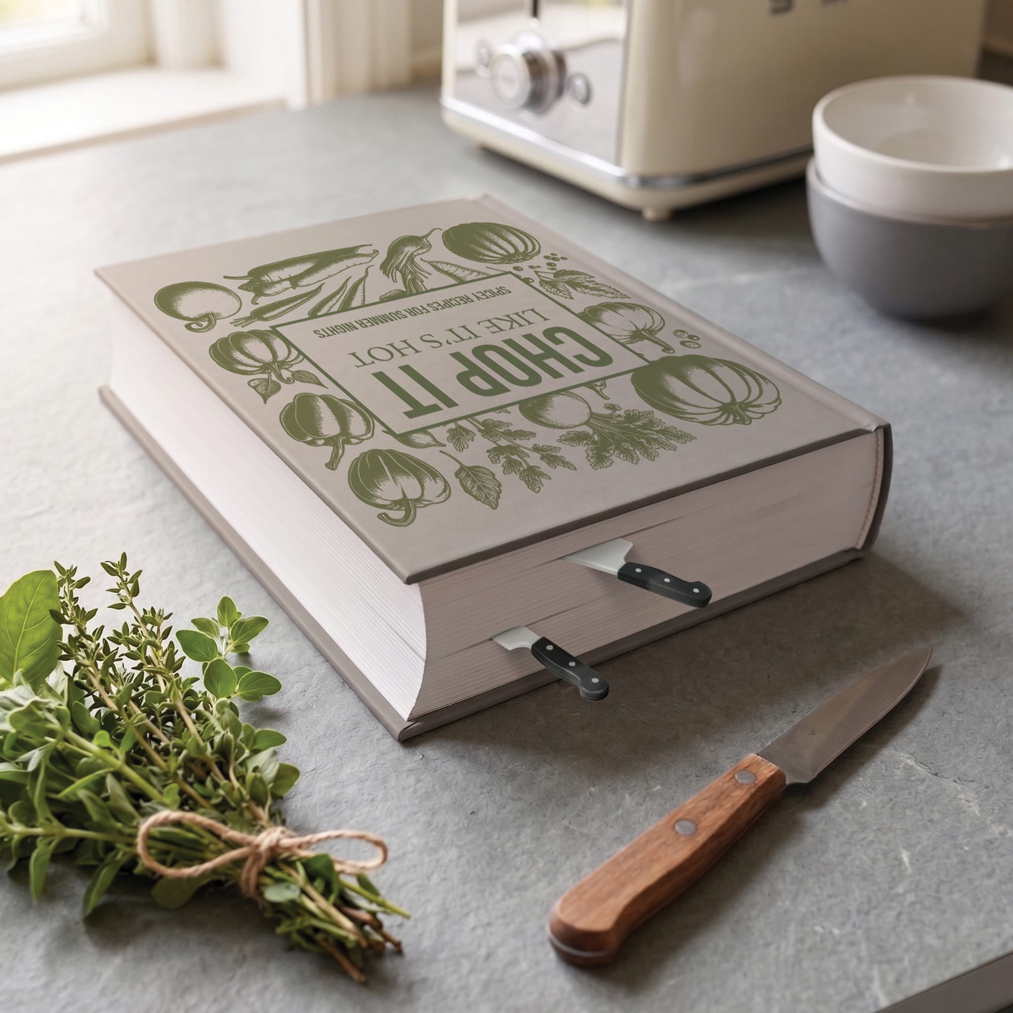 A decorative hardcover book with a green vegetable-themed cover design sits open on a gray countertop, with kitchen knife bookmarks inserted in the pages. A small bunch of fresh herbs and a wooden-handled knife are placed nearby, with a cream appliance and stacked bowls in the softly blurred background.
Cookmarks - mini chef knife bookmarks - make a great gift under $10 for home chefs, cooks, and cookbook lovers.
