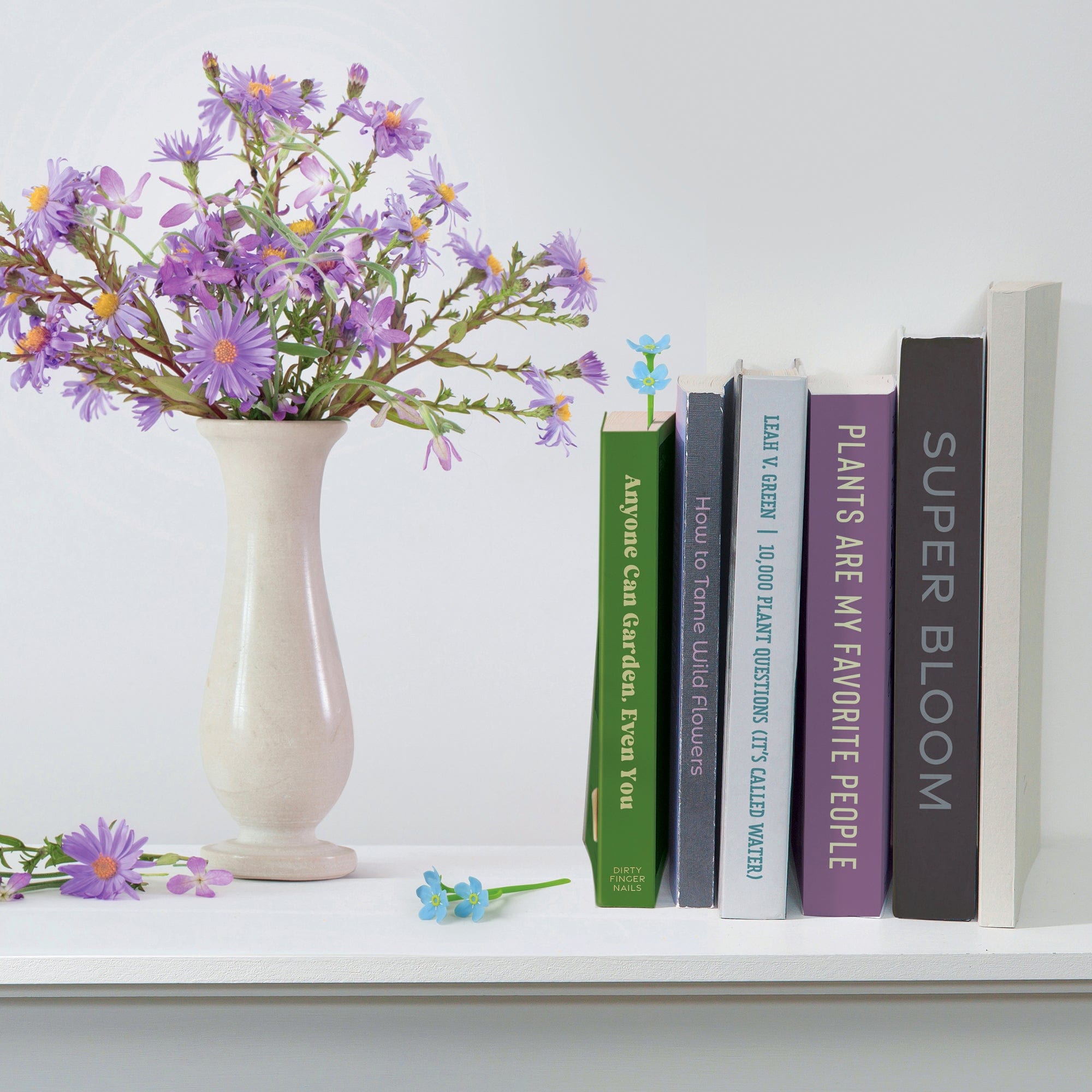 Vase with purple flowers next to books on a white surface. Book contains a bookmark that looks like a blue forget-me-not flower