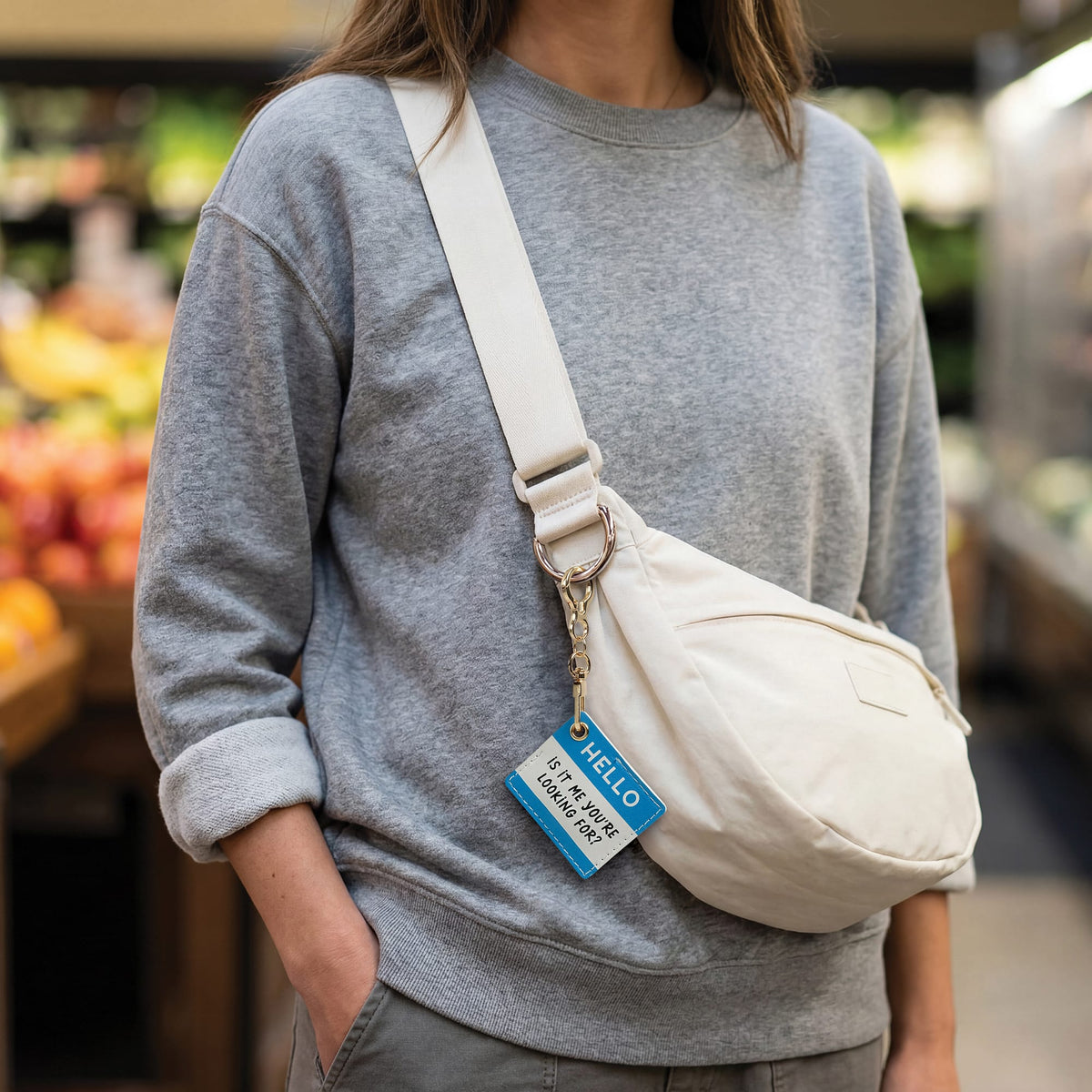 Person in a gray sweatshirt wearing a beige crossbody bag with a blue Hello tag on the strap.