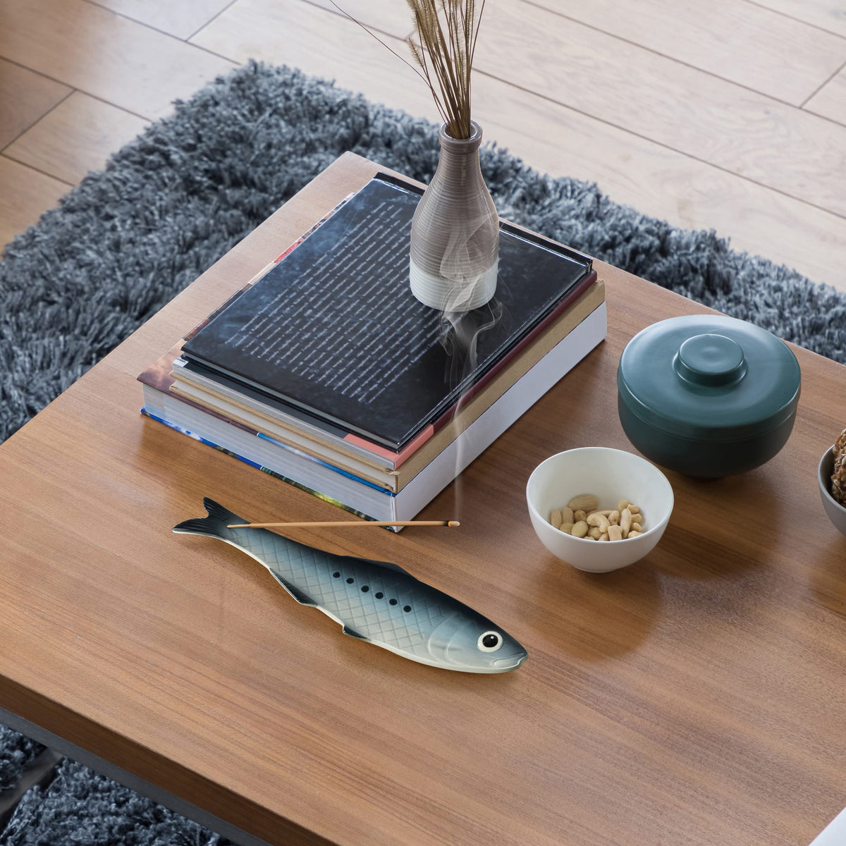 Cozy living room scene with a light gray ceramic vase holding incense sticks on a stack of books atop a wood coffee table near a gray rug.