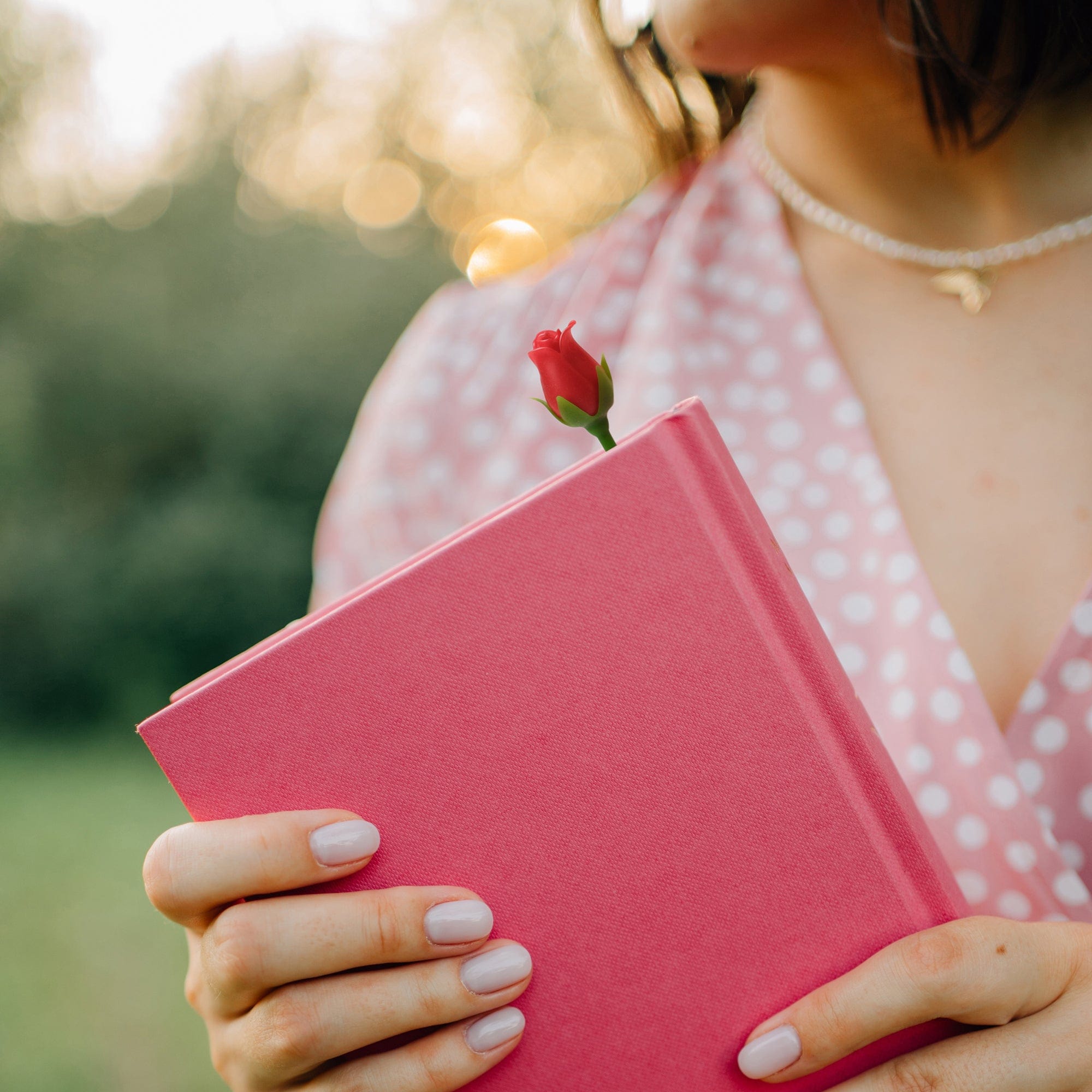 Person holding a pink book with a red flower bookmark on top, outdoors.