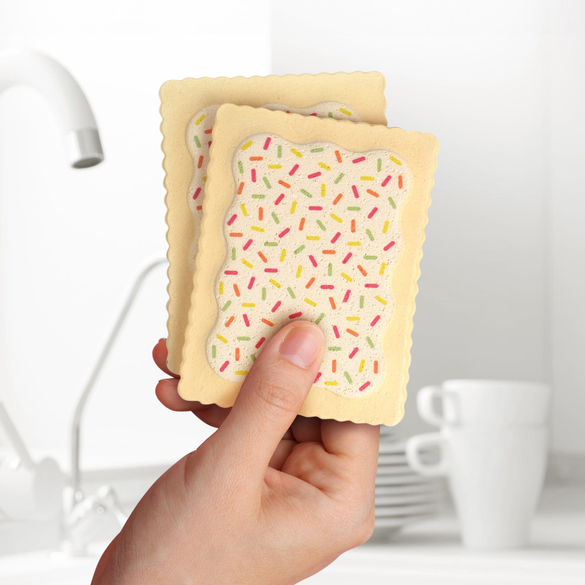 A hand holding Fred Slop Tarts Compressed Kitchen Sponges in front of a kitchen sink