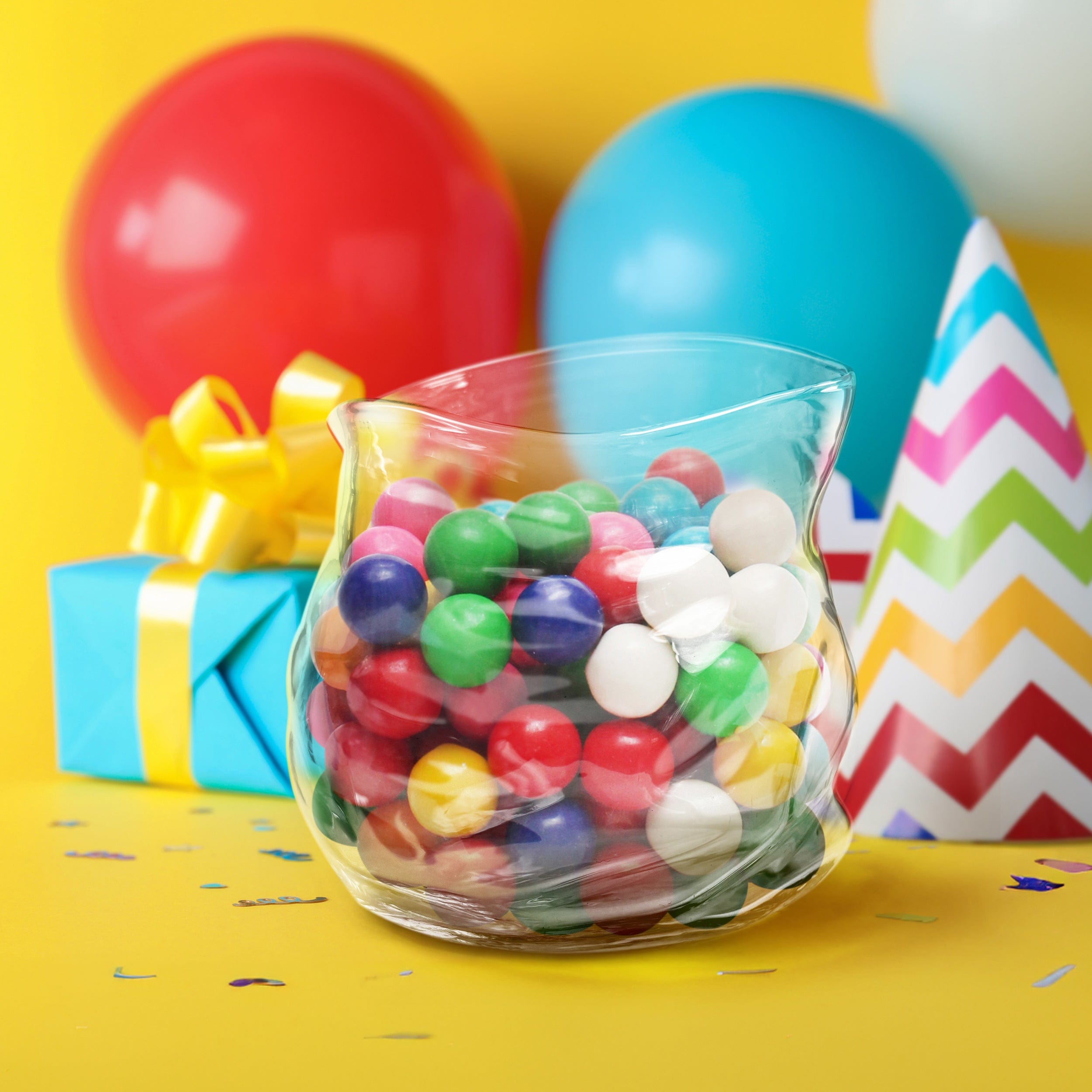 Glass vessel that looks like a ziploc zipper bag filled with colorful candies on a yellow background with balloons and a gift box.