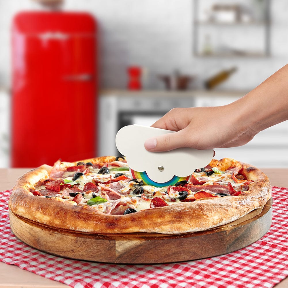 A woman's hand is cutting a pizza with a pizza cutter that looks like a cloud and rainbow. The handle is a cloud and the stainless steel blade is printed with a seven color rainbow. There is a kitchen in the background with a retro-looking red refrigerator.