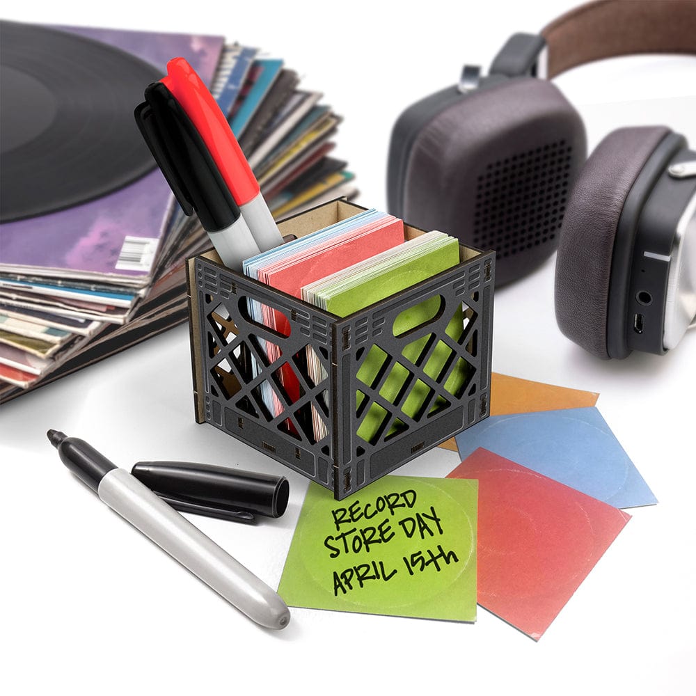 A small milk crate caddy for your desk. The crate is holding markers and notecards that look like vinyl album covers in 4 colors: orange, blue, green, and red. Behind the milk crate there is a stack of records and large headphones. There is a green notecard with handwriting on it that says " Record Store Day, April 15th"