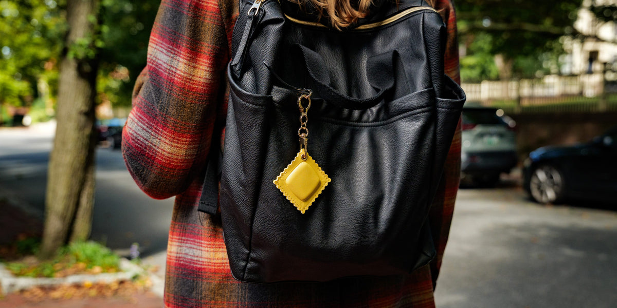 A person carries a black faux-leather backpack outdoors. A bright yellow square-shaped keychain or charm hangs from the backpack zipper with a gold-tone chain. A quirky, highly recommended holiday gift idea and budget-friendly stocking stuffer.