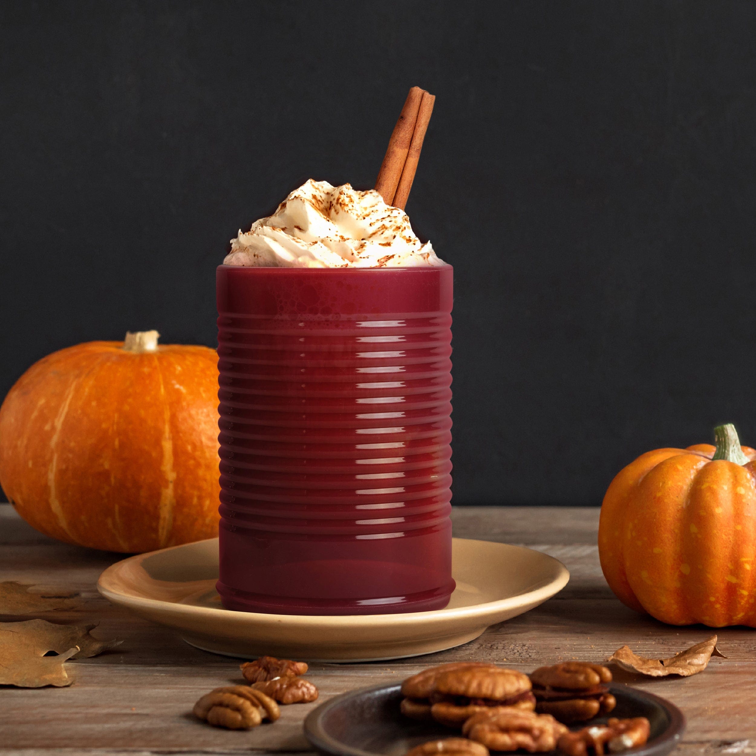 Pumpkin spice latte in a cranberry sauce cup with whipped cream and a cinnamon stick, surrounded by pumpkins and cookies on a wooden surface.