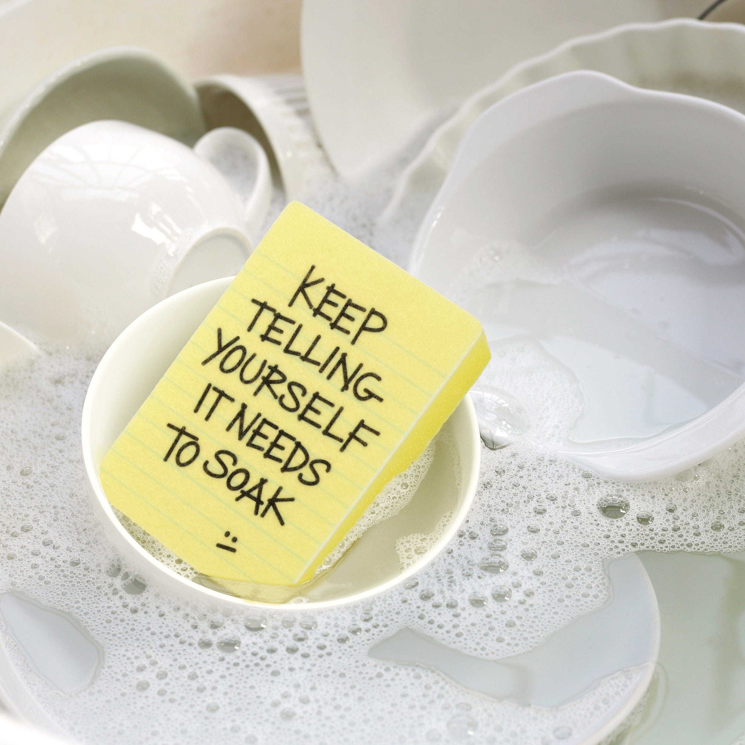 Yellow sticky note kitchen sponge with text on a white ceramic dish with soapy water.