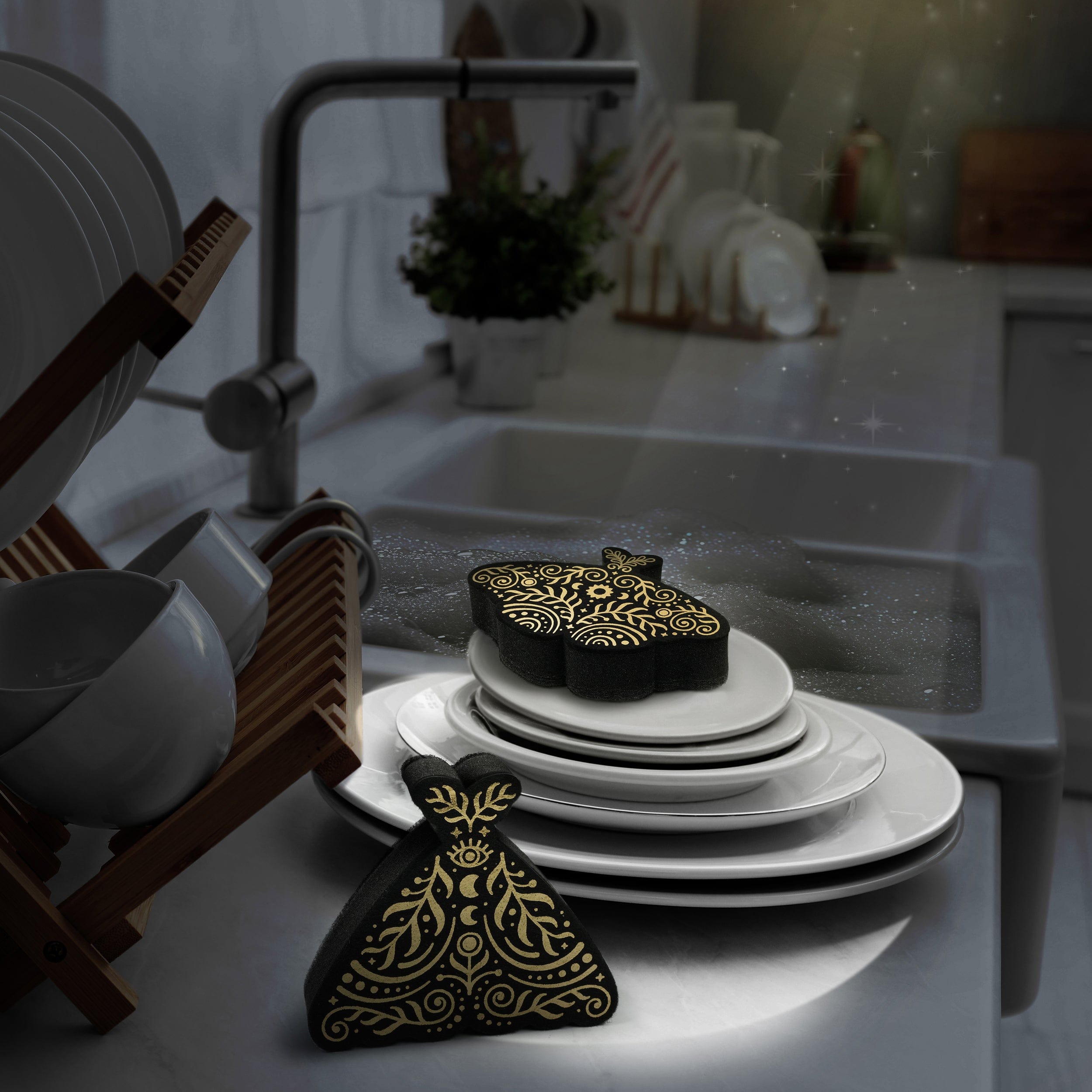 Black sponges that look like moths with abstract celestial details in metallic gold. The are sitting on white plates in a kitchen setting.