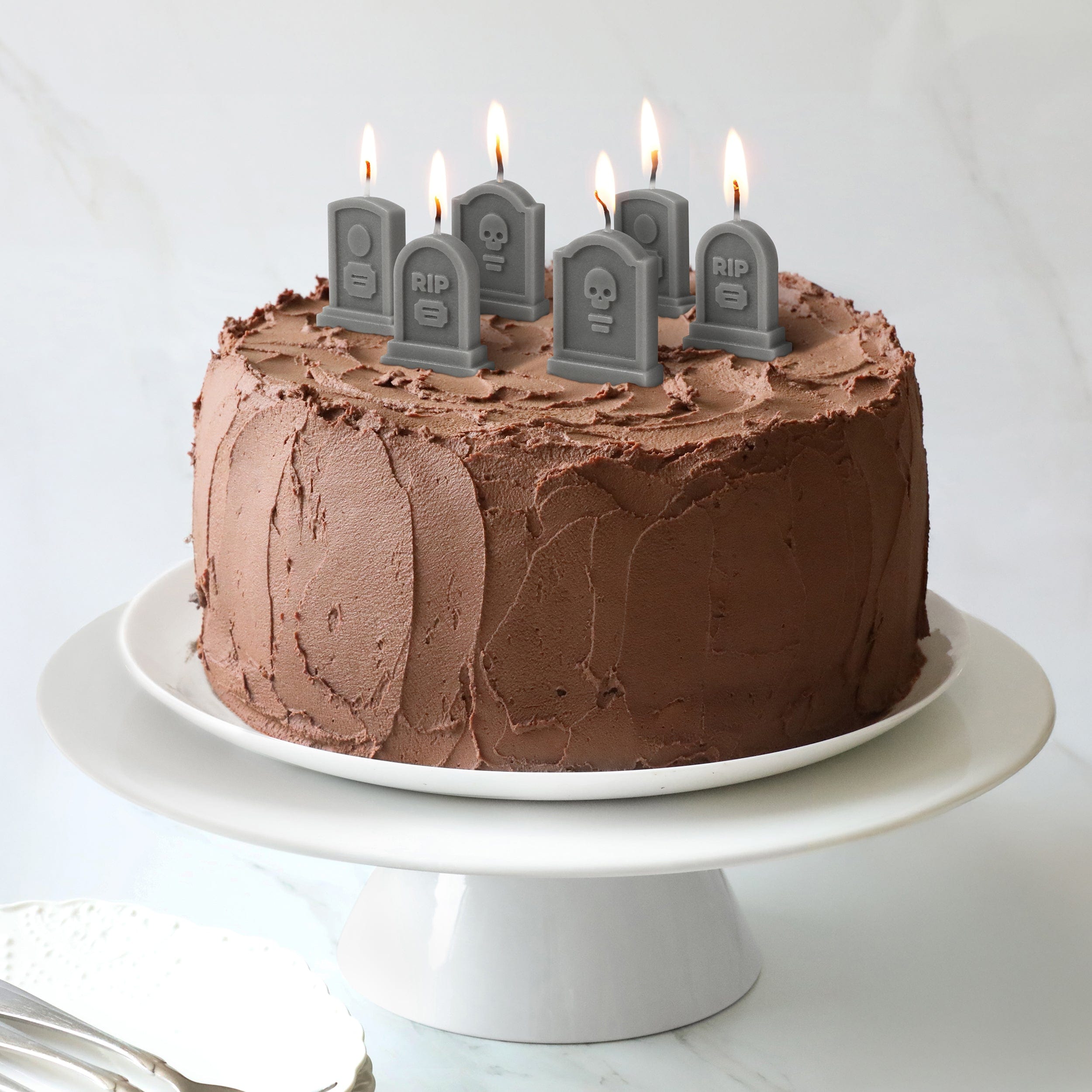 Chocolate cake with lit candles shaped like tombstones on a white plate.