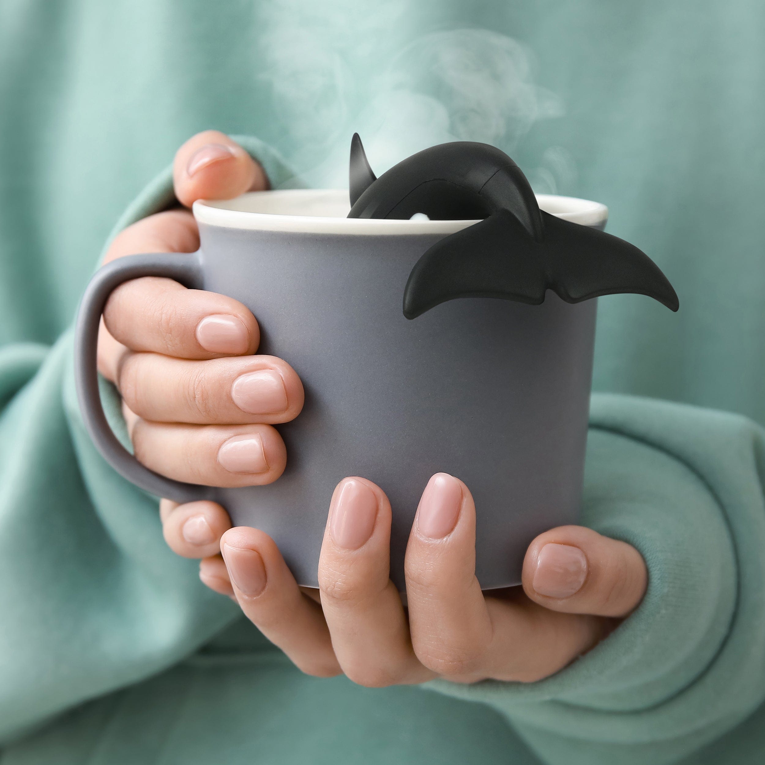 Person holding a gray mug with a black whale-shaped tea infuser against a green background