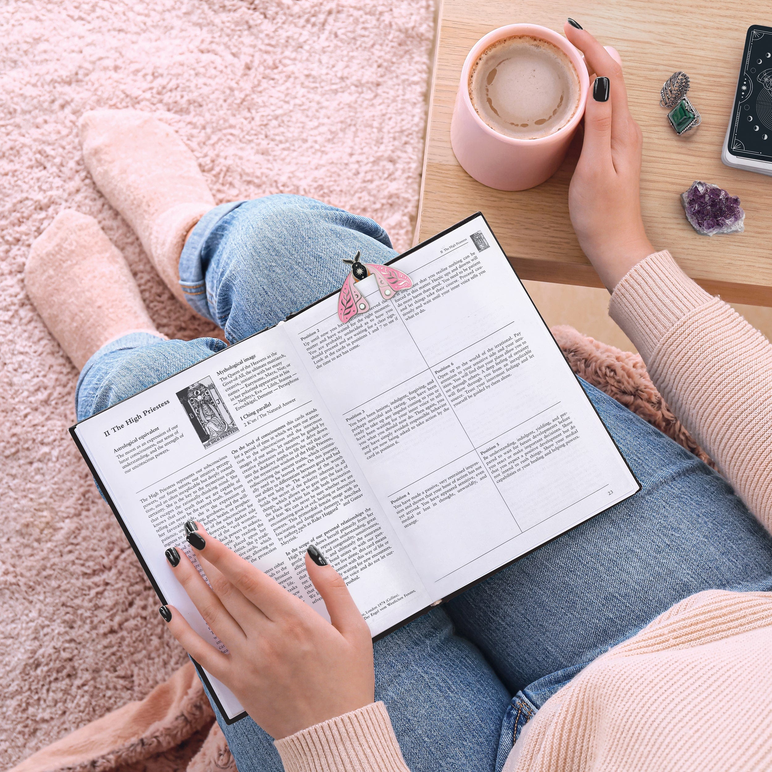 Person reading a book marked with a moth bookmark and a cup of coffee on a wooden table