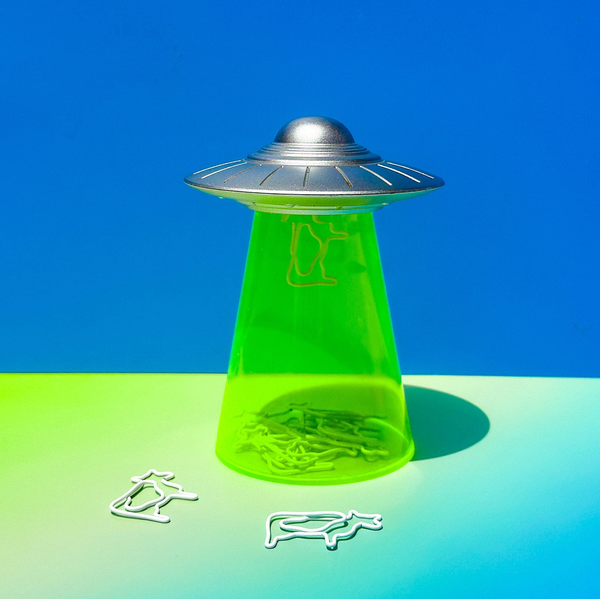 Green UFO-shaped container with silver top on a blue and green gradient background. Cow paperclips