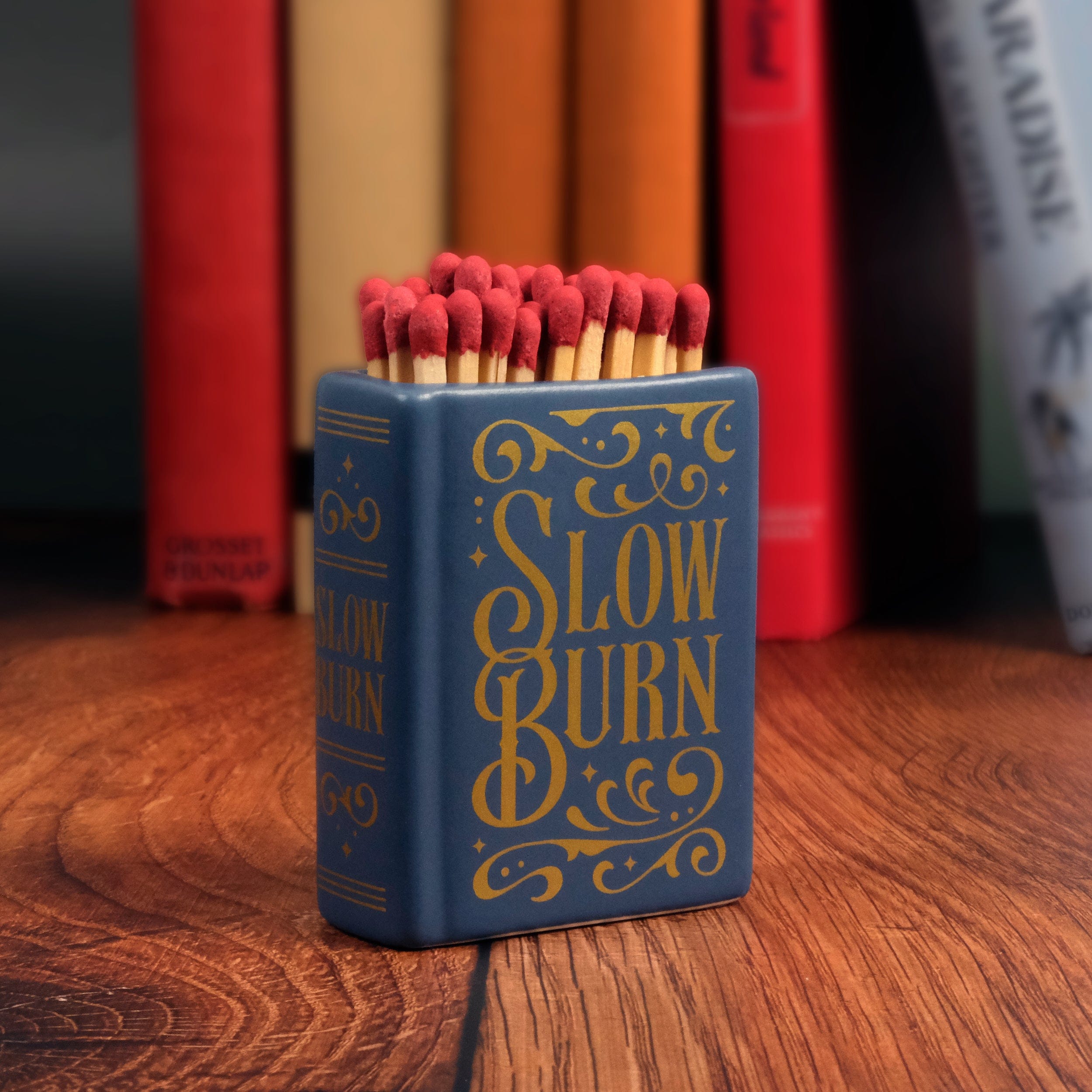 Book Lover Gift under $20. Ceramic match holder labeled 'Slow Burn' with matches on a wooden surface, books in the background.