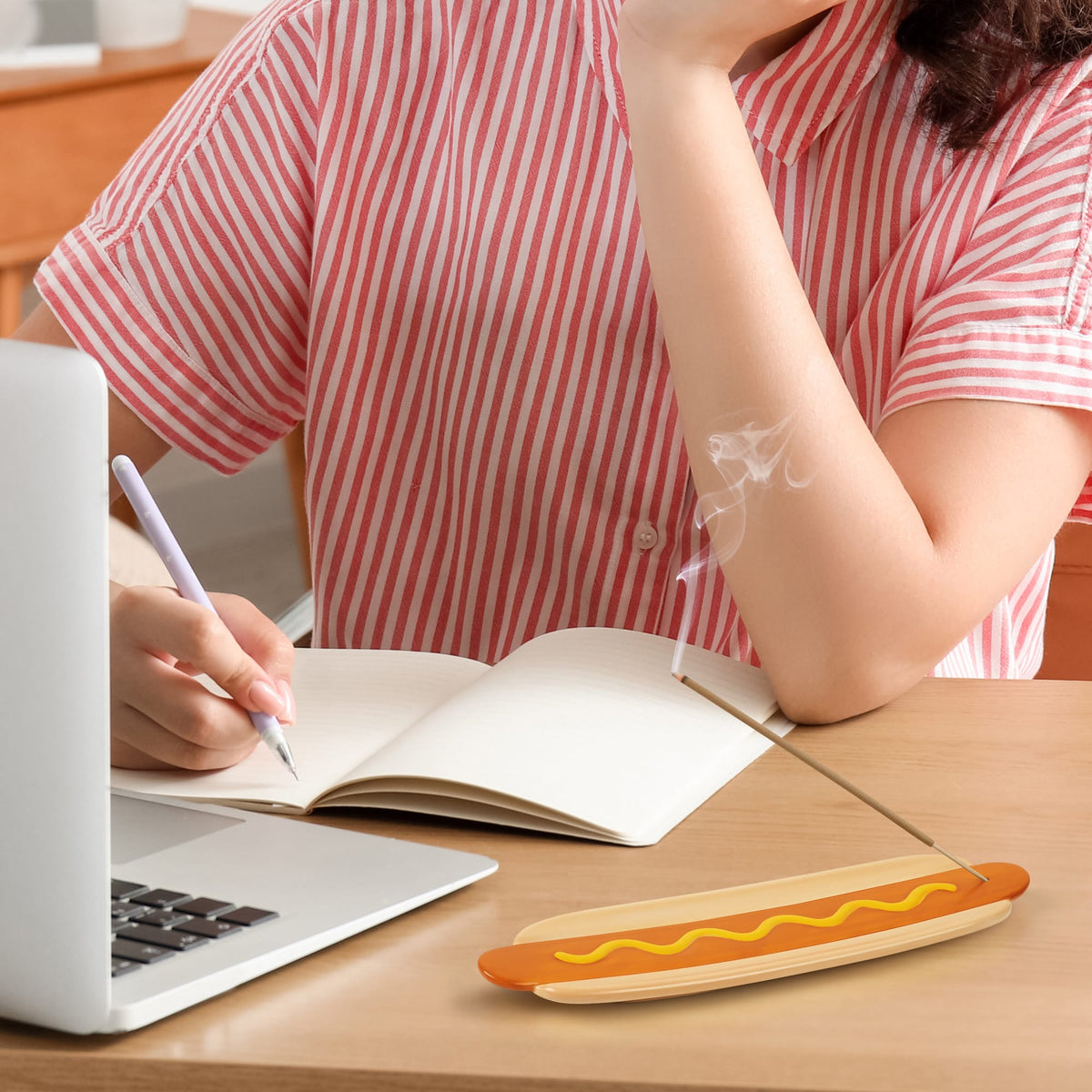 Person in a striped shirt writes in a notebook with a Frank Incense incense stick nearby on the desk.