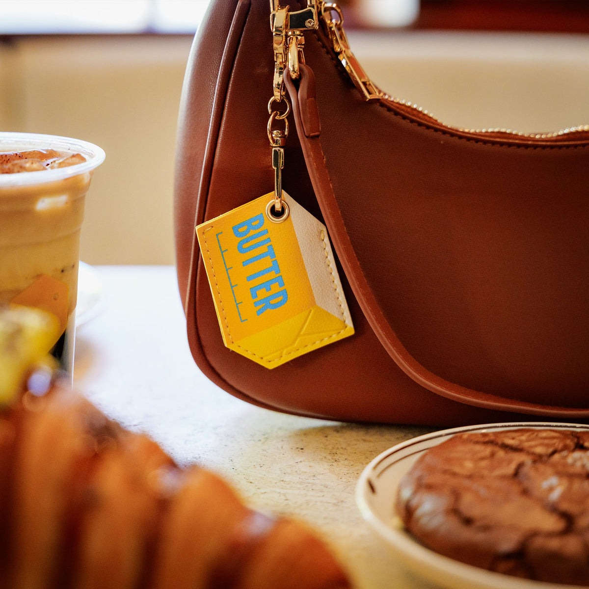 A brown leather shoulder bag with a bright yellow tag featuring the blue 'BUTTER' logo, styled on a cafe table next to a drink and pastries. A fun and functional holiday gift idea for foodies.