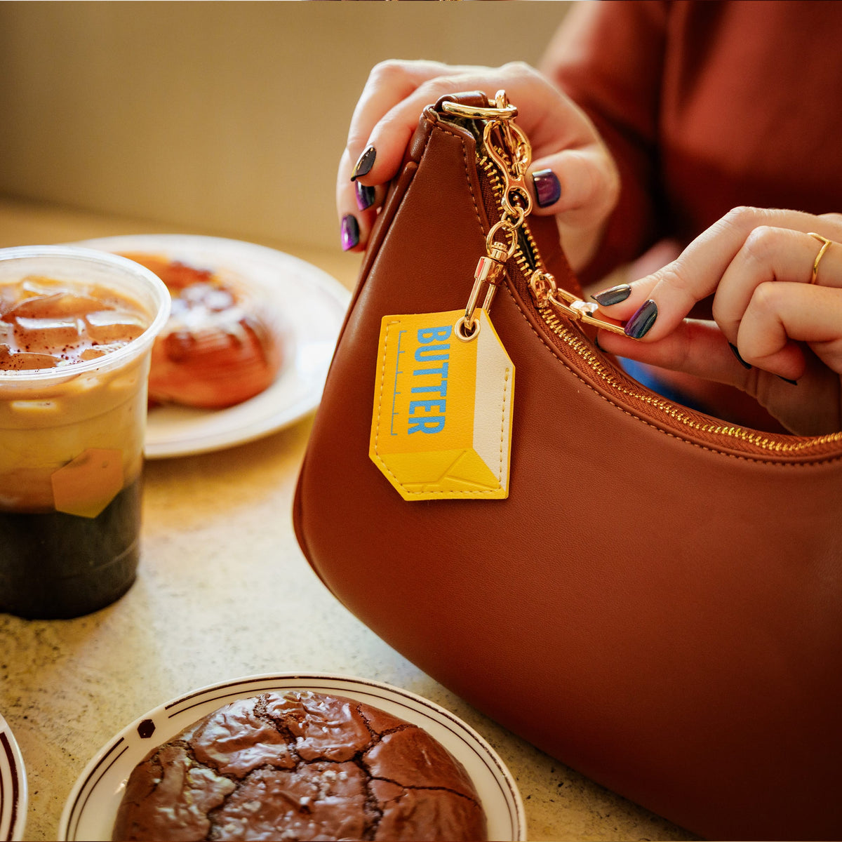 A person's hands with purple and black nail polish are shown placing a yellow keychain charm into a brown leather handbag. The charm features the word 'BUTTER' in blue text on a bright yellow background, attached to a gold-tone chain. A fun and functional holiday gift idea for foodies, friends, teachers, and teens.