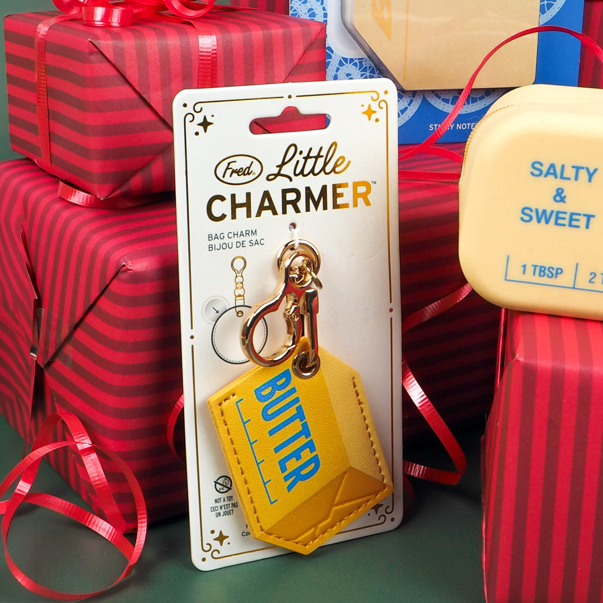 A yellow bag charm shaped like a butter block with blue 'BUTTER' text, attached to a gold keychain and displayed on a white 'Fred Little Charmer' backing card. The card is photographed in a festive setting surrounded by red wrapped gift boxes and ribbons.