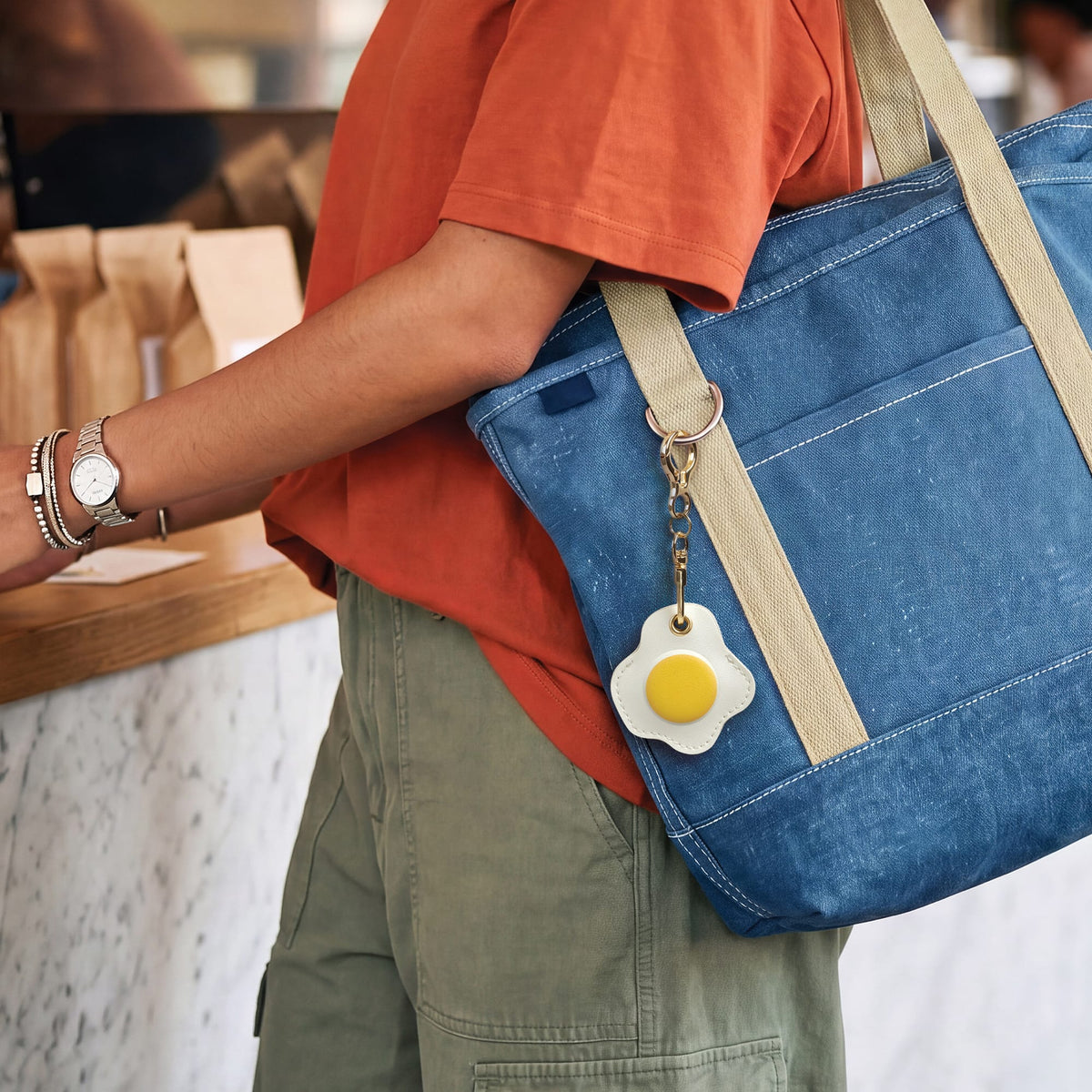 Person wearing an orange shirt carrying a blue bag with beige straps, accented with the fried egg bag charm. A trendy everyday holiday gift and fun, affordable stocking stuffer $10 and under.