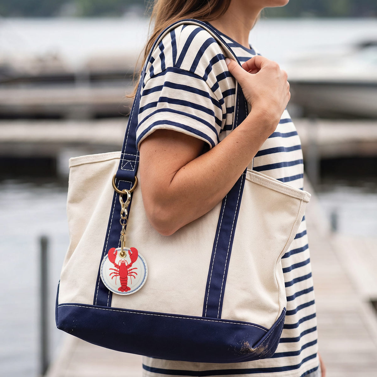 Person in a nautical striped shirt carrying a beige canvas tote bag accented with the lobster bag charm. A clever, budget-friendly tech accessory holiday gift and useful stocking stuffer.