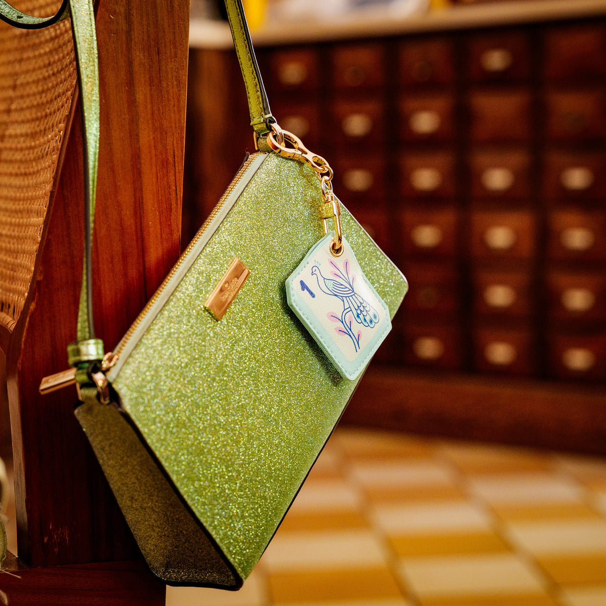 A green glittery shoulder bag with gold hardware and a decorative mahjong bag charm. Mahjong bird tile bag charm clipped to a purse strap. A stylish, novelty holiday gift for board game enthusiasts and an easy stocking stuffer $10 and under.