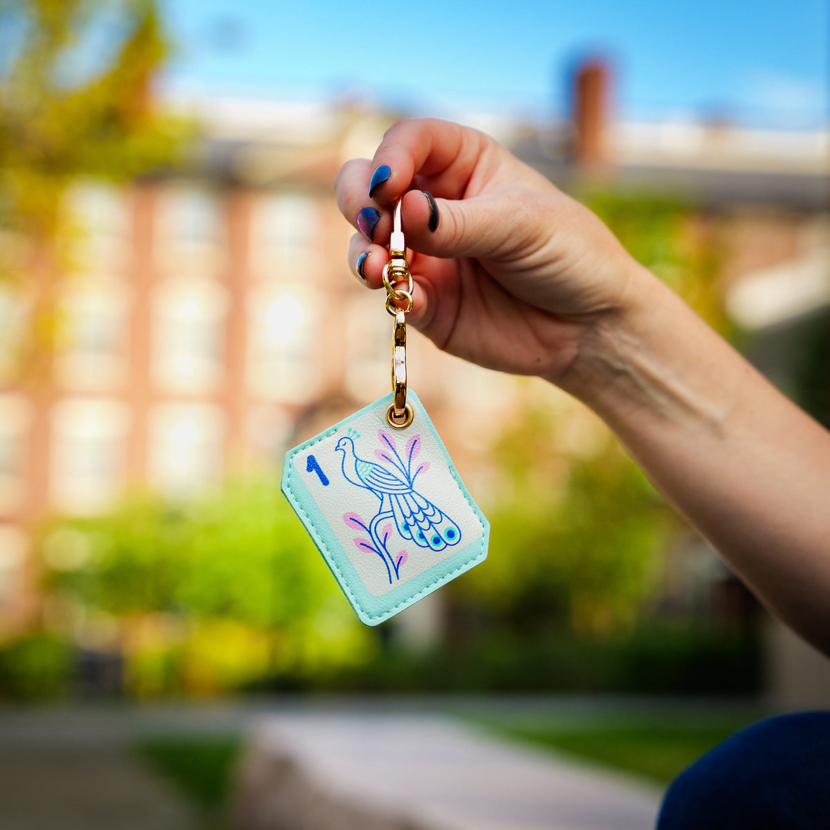 Person holding the Mahjong bird tile bag charm, showing its compact size and functional design. A top-rated practical stocking stuffer and fun holiday gift for tabletop gamers. Blurred outdoor campus background.