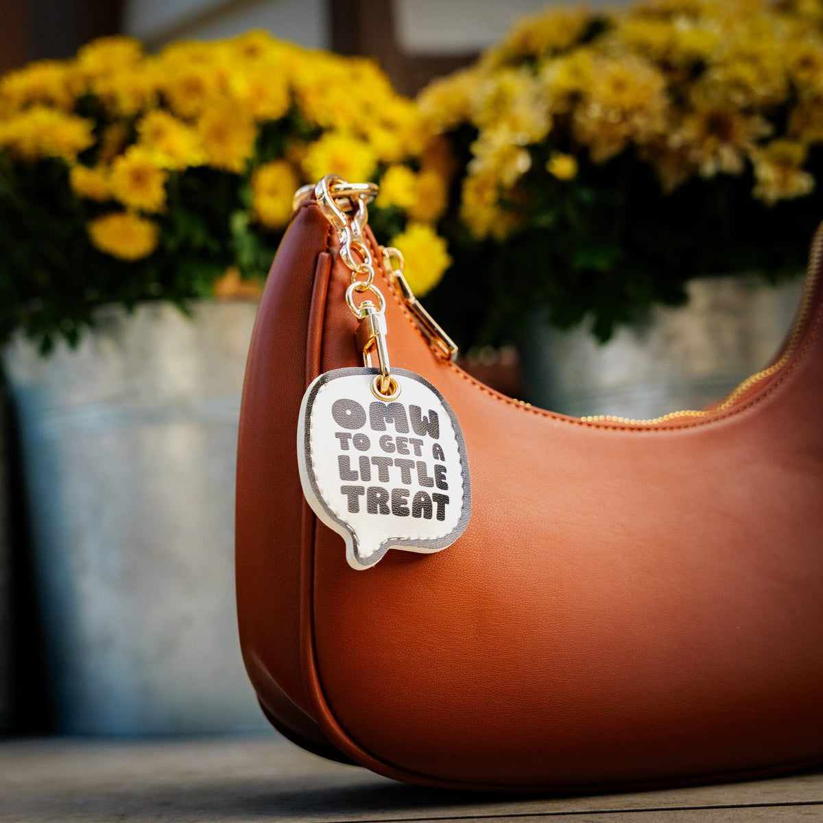 A brown leather handbag with a gold zipper, featuring a white speech-bubble-shaped keychain charm that reads 'OMW TO GET A LITTLE TREAT' in dark gray text. The background is a soft-focus outdoor setting with yellow flowers in metal planters.