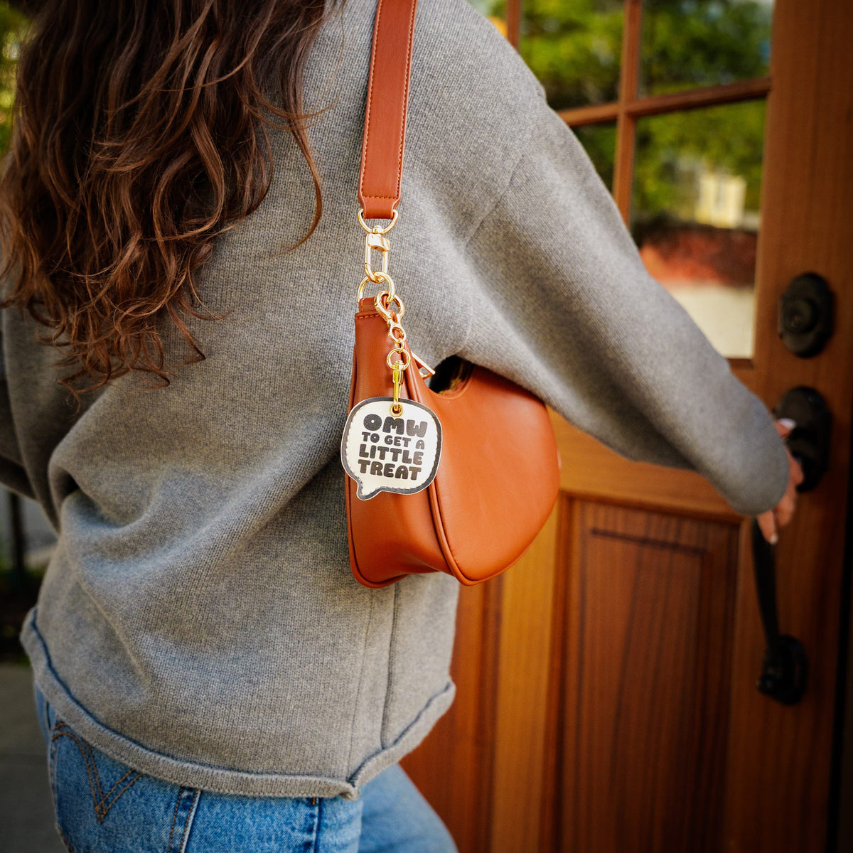 A woman wearing a gray sweatshirt and blue jeans carries a small brown leather shoulder bag with a gold chain strap and a white speech-bubble charm that reads 'OMW TO GET A LITTLE TREAT'. She is standing in front of a wooden door. A trendy, highly rated holiday gift idea and budget-friendly stocking stuffer for friends.