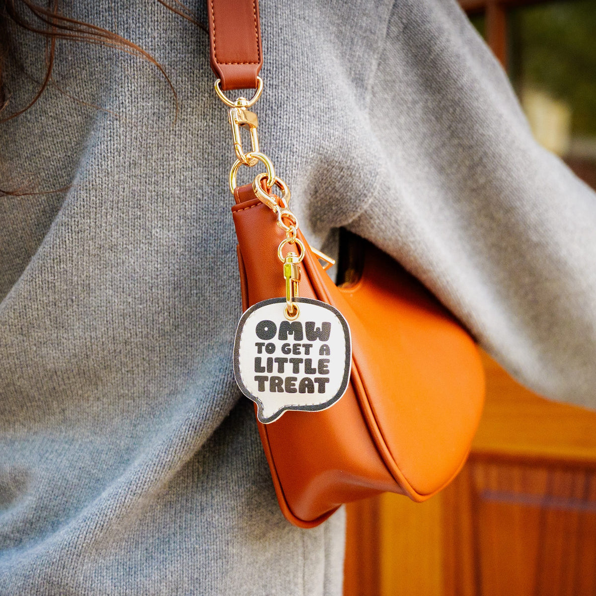 Close-up of the white circular bag charm with bold text reading 'OMW TO GET A LITTLE TREAT'. A top-rated, relatable novelty holiday gift and practical everyday carry item under $10.