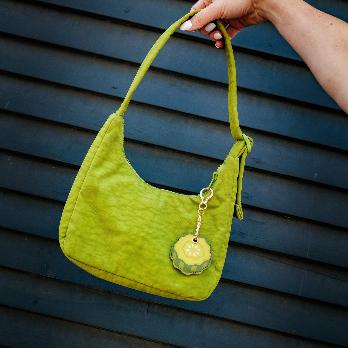 A lime green textured fabric shoulder bag with a matching strap and a decorative pickle-shaped charm, held by a hand against a dark blue wooden slat wall.