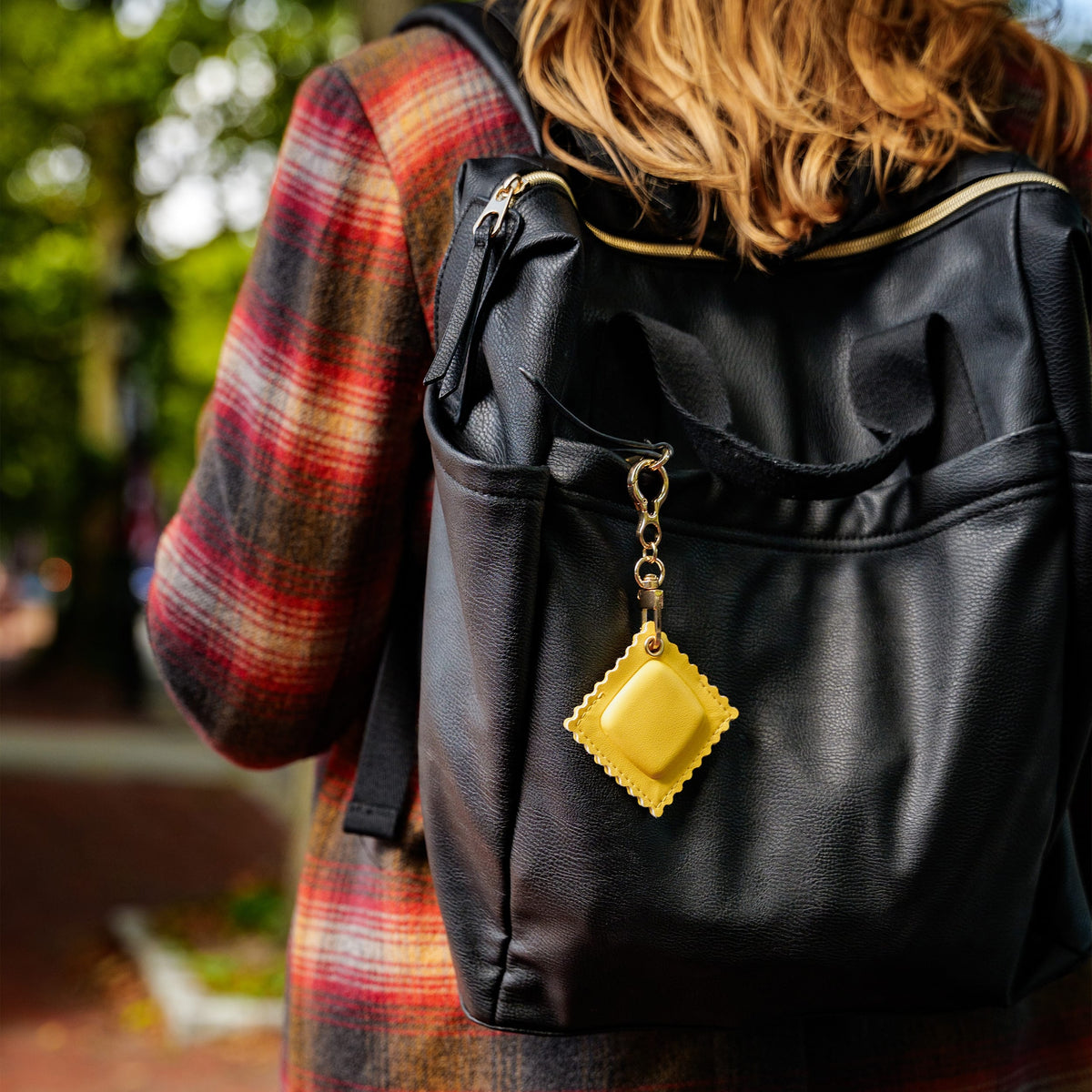 A person wearing a red and black plaid coat carries a black faux-leather backpack outdoors. A bright yellow square-shaped keychain or charm hangs from the backpack zipper with a gold-tone chain. A quirky, highly recommended holiday gift idea and budget-friendly stocking stuffer.