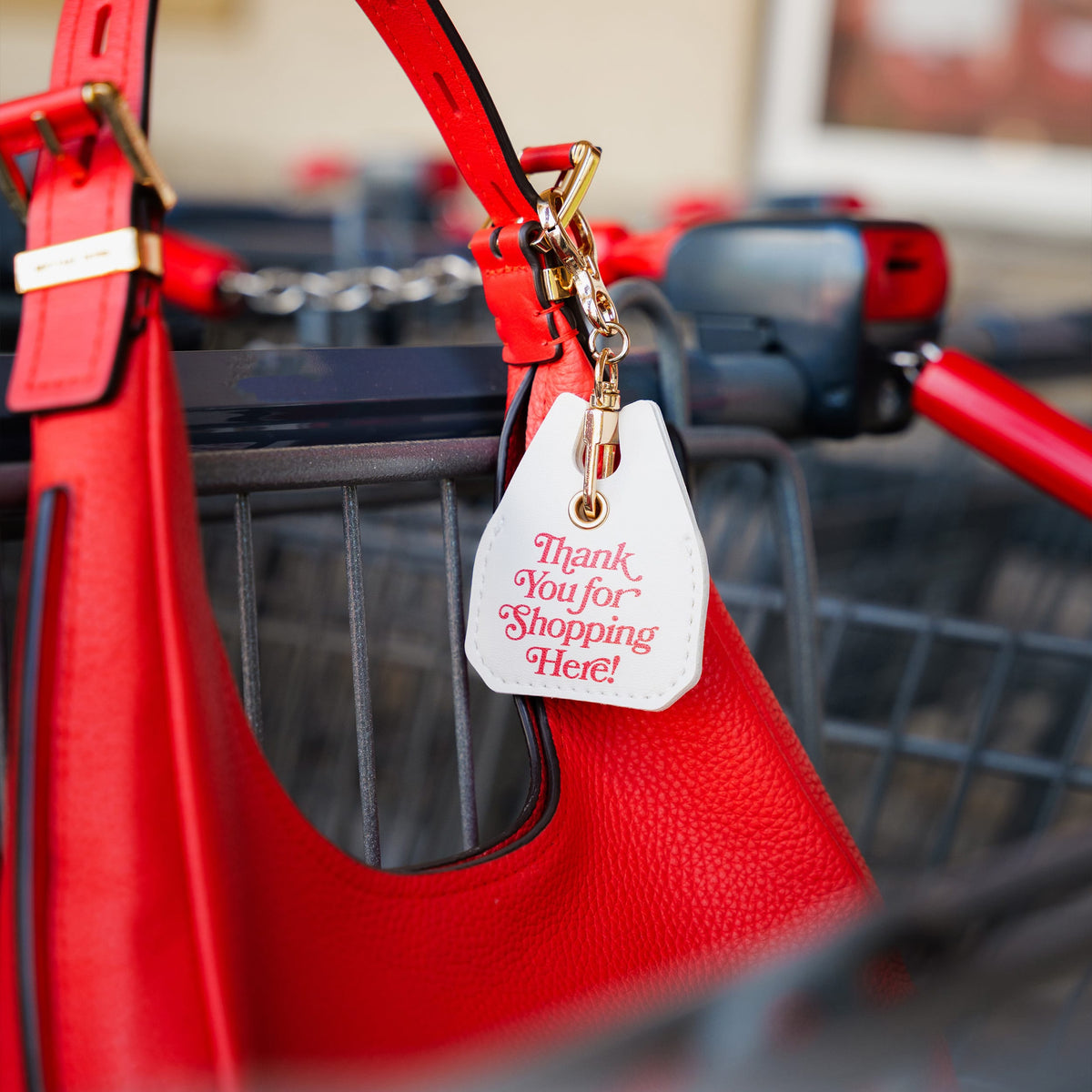 A bright red textured leather shopping bag hanging in a metal shopping cart, with a white hanging tag that reads 'Thank You for Shopping Here!' in red script. A clever, highly useful holiday gift and top stocking stuffer idea for grocery shoppers.