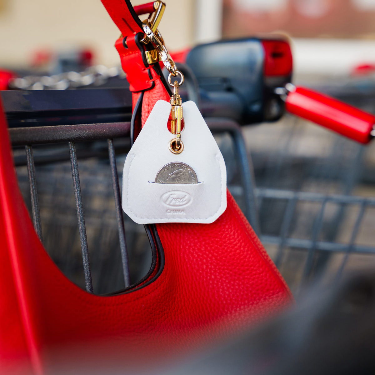A white leather coin purse keychain with a gold clasp and a visible US quarter, hanging from a red shopping bag next to a shopping cart. Attached to a shopping cart, demonstrating the built-in emergency quarter slot. A clever, highly useful holiday gift and top stocking stuffer idea for grocery shoppers.