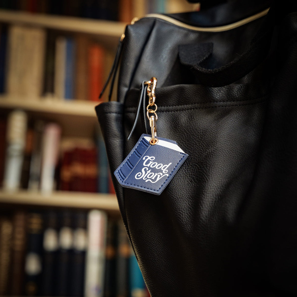 A close-up of a black leather tote bag with a blue leather keychain tag that reads 'Good Story' in white script, hanging from a gold-tone ring in front of a blurred bookshelf background.

A practical and easy stocking stuffer $10 and under. Great gift for book lovers, readers, students, teachers, and teens.