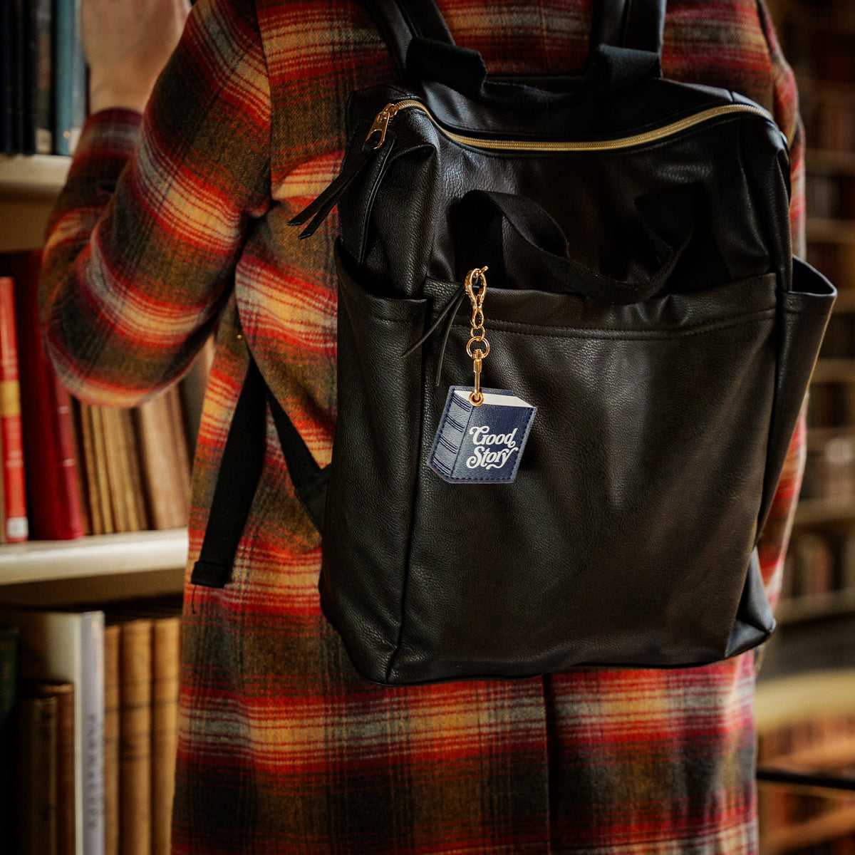 A black faux-leather backpack with a gold zipper and a hanging 'Good Story' tag, worn by a person in a red and orange plaid coat in front of a bookshelf.