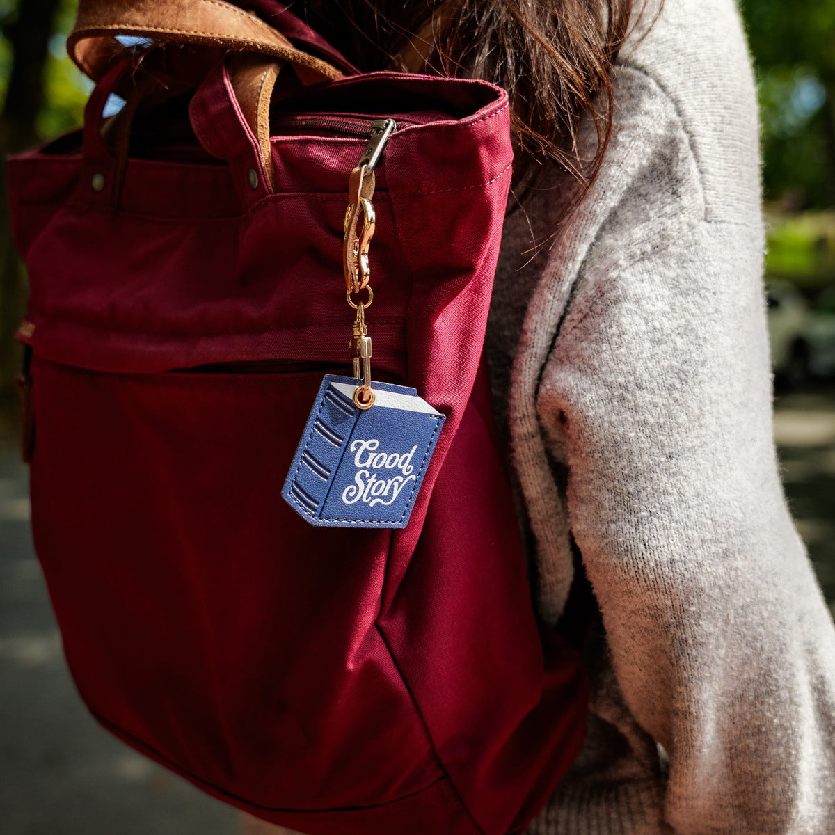 A close-up of a person wearing a burgundy canvas tote bag with brown leather handles and a blue book-shaped keychain charm that reads 'Good Story'.