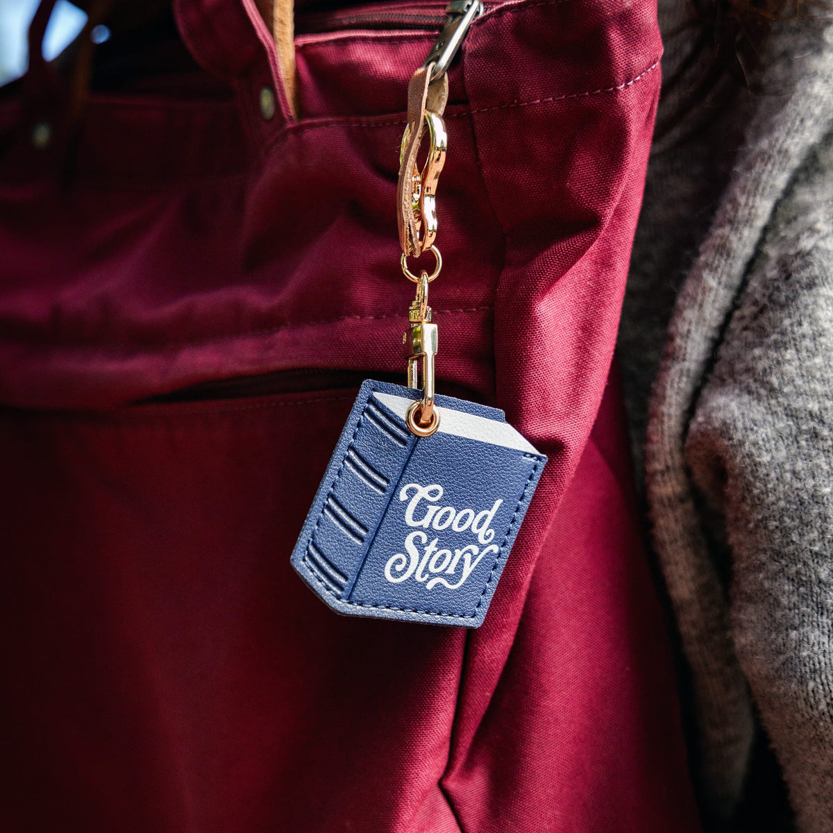 A close-up of a navy blue faux-leather keychain shaped like a book, featuring the words 'Good Story' in white script, attached to a gold key ring and clasp. The keychain is shown hanging from a burgundy nylon bag next to a gray wool jacket. A stylish accessory and great gift for book lovers.