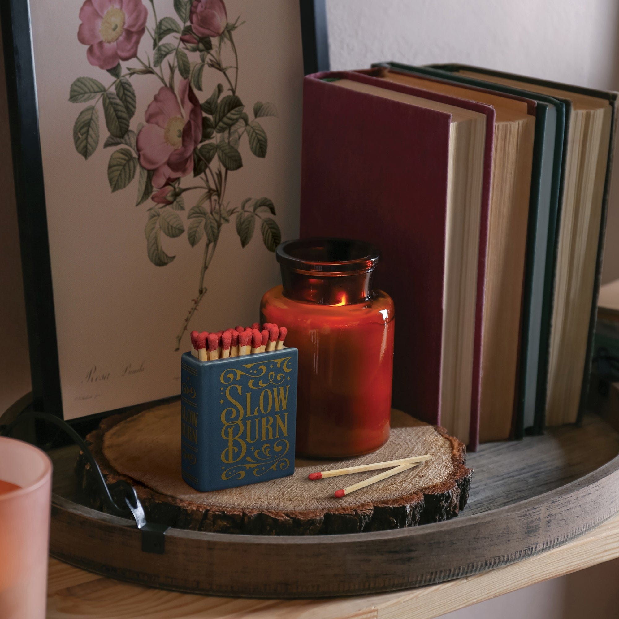 Candle, matches, and books on a wooden surface with a floral painting in the background. Unique literary gift and reading nook decor